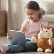 Curious little child girl having fun using digital tablet alone embracing toy sitting on floor, happy preschool smart kid playing with computer looking at screen watching cartoons online at home